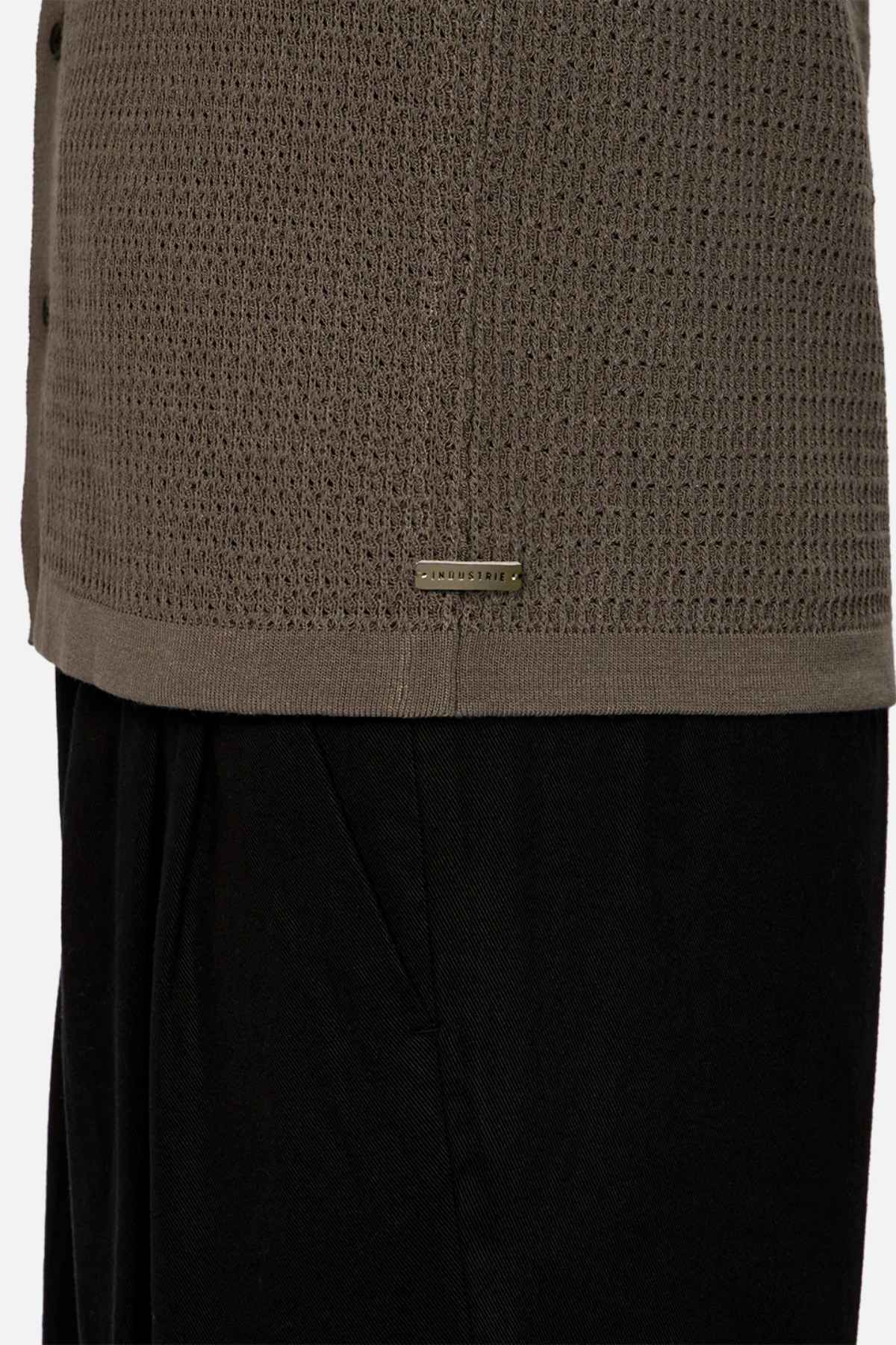 A close-up showcases someone wearing the textured olive green Industrie The Sevona Short Sleeve Shirt, an ideal addition to a summer wardrobe with its relaxed fit and subtle rectangular tag near the hem. It's stylishly paired with black pants against a plain background.