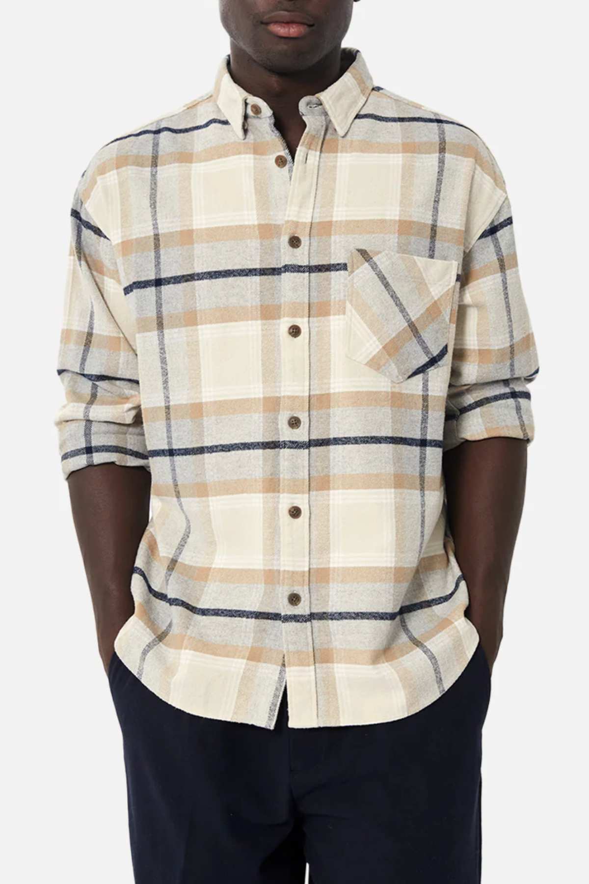 A person wears the Industrie The Somerville Shirt, a classic long-sleeved beige/navy/light orange plaid shirt with rolled-up sleeves. The left chest pocket subtly displays the Industrie branding. With hands in dark pants pockets, they stand against a plain white backdrop.