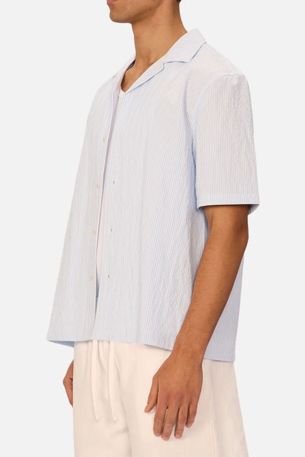 A man stands in profile wearing the Industrie The Sorrento Short Sleeve Shirt, a light blue and white pinstripe cotton shirt, paired with beige drawstring pants against a plain background.