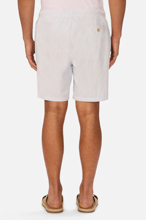 A person stands facing away, wearing Industrie The Sorrento Short in light blue and white pinstripes with an elastic waistband, a single buttoned back pocket, tan sandals, and a white shirt.