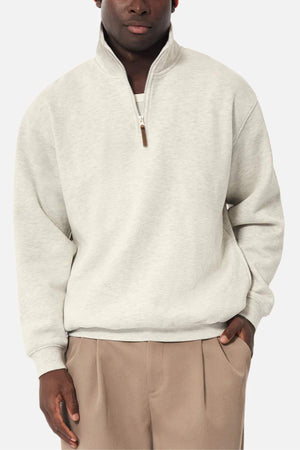 A man wearing an Industrie The Staunton Tracktop in light gray and tan pleated trousers stands with one hand in his pocket against a plain white background.