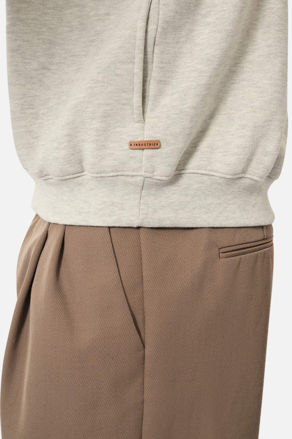 A close-up of a person wearing the Industrie The Staunton Tracktop in light grey, fleece-lined, with a small brown tag, paired with high-waisted brown pleated trousers featuring a welt pocket.