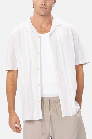 Someone is wearing the Industrie Striped Magna Short Sleeve Shirt in white and light gray over a white tank top, paired with light beige drawstring pants. The breathable fabric ensures comfort, while the plain white background adds to the simplicity of the style.