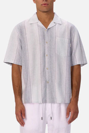 A man wears the Industrie The Sunset Short Sleeve Shirt, a men's casual shirt featuring grey and white vertical stripes and a chest pocket, paired with white pants against a plain light background.