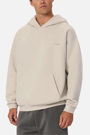 A man wears the Industrie The Tech Del Sur Hood in light beige, featuring a front pocket and subtle chest logo, paired with dark gray athletic pants, standing against a plain white background.