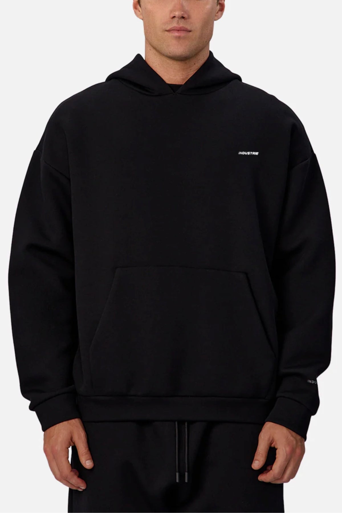 Someone wears the Industrie The Tech Del Sur Hoodie—plain black with a small white logo on the left chest. This athletic hoodie has a front pocket, loose fit, and is shown against a light gray background.