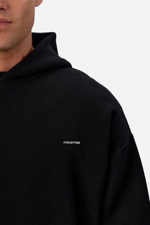 A person wears the Industrie The Tech Del Sur Hoodie—plain black with "INDUSTRIE" in small white chest lettering. Only their lower face and upper body are shown, set against a plain background.