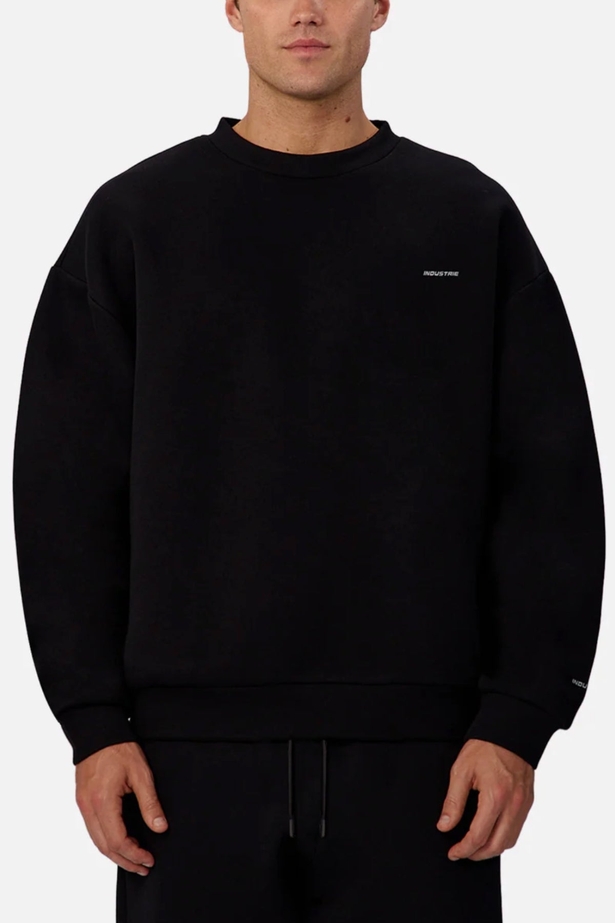 A person wears the loose-fit black Industrie The Tech Del Sur Sweat, featuring small white text on the left chest and wrist, with black pants, standing against a plain light background.