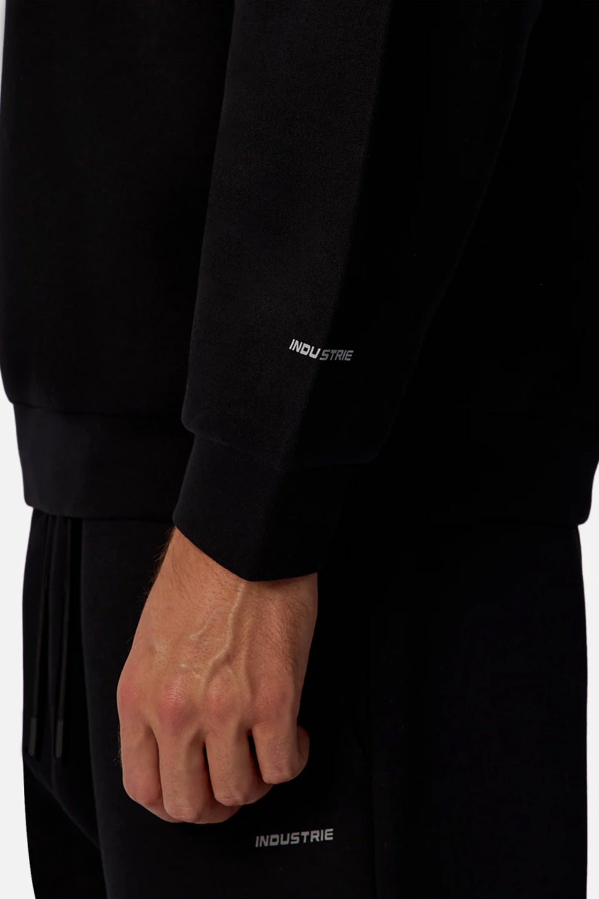A close-up of a hand and arm wearing the black Industrie The Tech Del Sur Sweat, featuring "INDUSTRIE" in small white letters on the sleeve and pant leg.