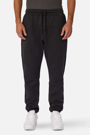 Dressed in Industrie The Tech Del Sur Track Pant and white sneakers, an individual stands against a plain white background. The sweatpants, made from athletic material, have an elastic drawstring waistband. Only the lower half is visible, paired with a dark upper garment.