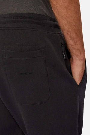 Close-up of someone wearing Industrie The Tech Del Sur Track Pant with dark athletic material, a visible back pocket, and their right hand in the front pocket. The fabric, featuring an elastic waistband, looks soft against a neutral background.