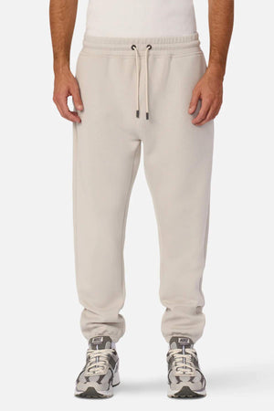A person is wearing light beige jogger pants with an elastic waistband and cuffs, resembling the Industrie The Tech Del Sur Track Pant, paired with a white sleeveless top and gray and white sneakers. Only the lower half of the body is visible, showcasing the clothing and shoes.