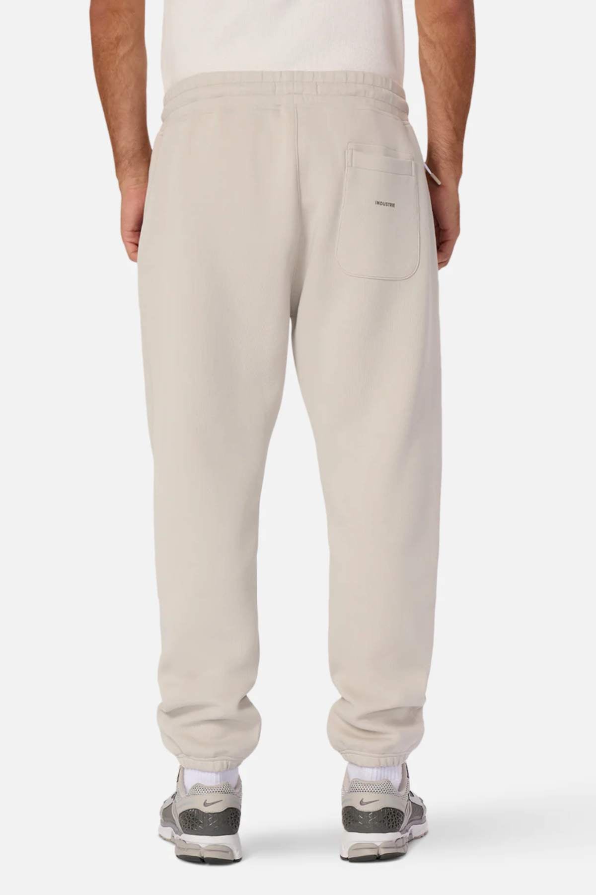 A person stands with their back facing the camera, wearing light beige Industrie The Tech Del Sur Track Pant joggers made from athletic material with an elastic waistband and a single back pocket. They are also sporting gray and green athletic shoes. The background is plain white.