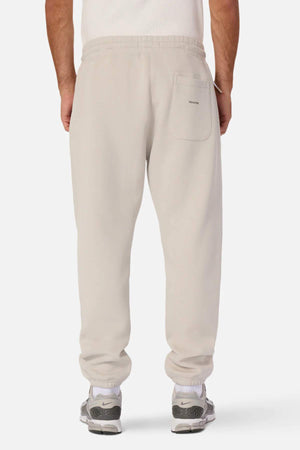 A person stands with their back facing the camera, wearing light beige Industrie The Tech Del Sur Track Pant joggers made from athletic material with an elastic waistband and a single back pocket. They are also sporting gray and green athletic shoes. The background is plain white.