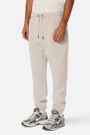 A person is wearing Industrie The Tech Del Sur Track Pants made from athletic material, featuring a light beige color with an elastic waistband and drawstring. They are also sporting light grey and white athletic sneakers with white socks. The person's upper body is out of the frame, and the background is plain white.