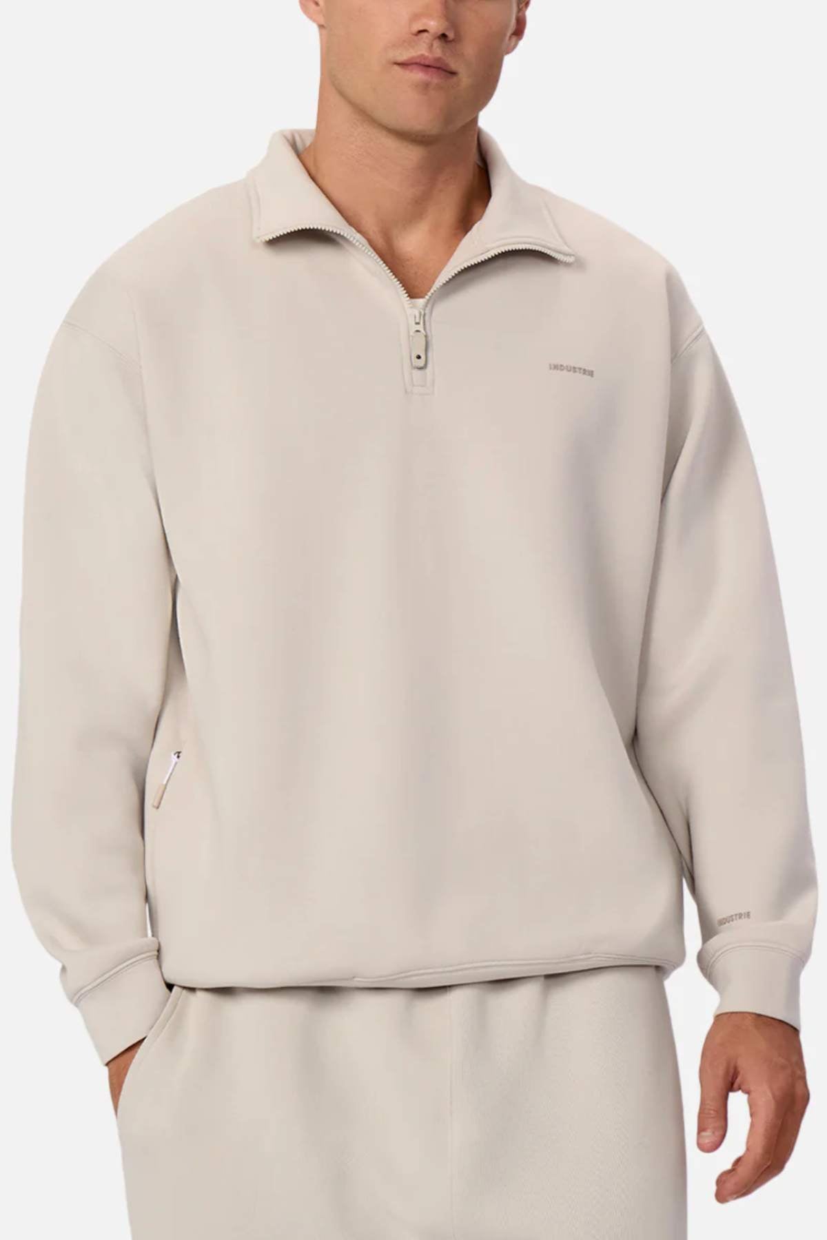 Someone is dressed in a light beige Industrie The Tech Staunton Track Top with matching pants, made of athletic fabric. It boasts a standing collar, zip-up side pockets, and subtle chest branding, creating a comfortable and casual look against a plain background.