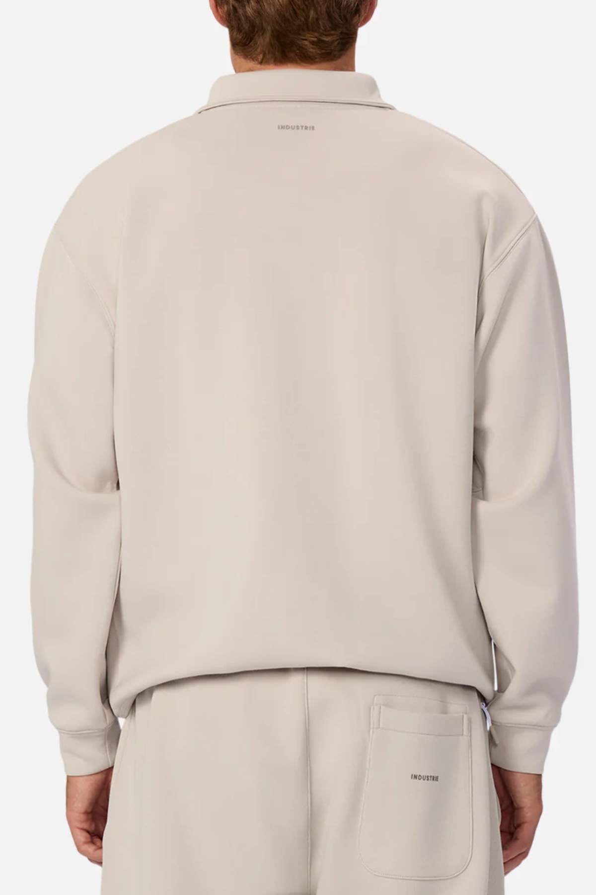 A person is seen from the back in light beige long-sleeve "Industrie The Tech Staunton Track Top" with matching pants. The athletic set features minimal branding with "Industry" on the upper back and pants' pocket, and zip-up side pockets for functionality, posed against a white background.