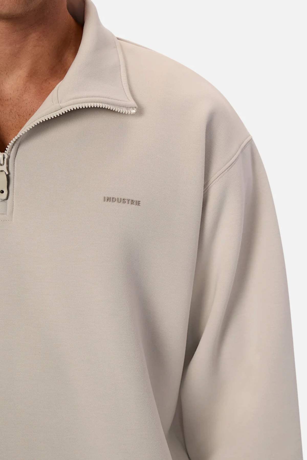 A person wearing the Industrie The Tech Staunton Track Top in light gray with "INDUSTRIE" embroidered on the chest, reveals part of their neck and shoulder with an unzipped quarter-zip collar. This athletic pullover includes zip-up side pockets against a plain white background.