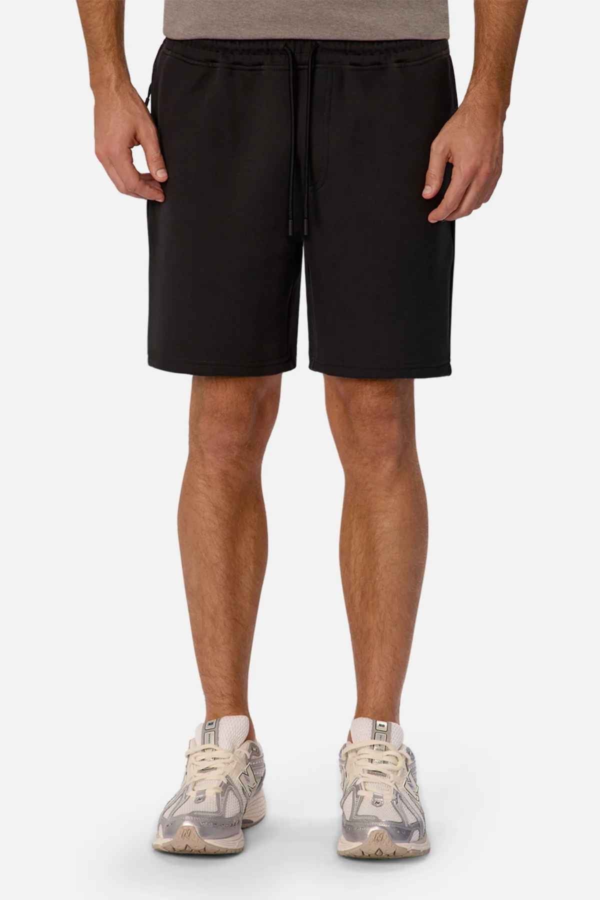 A person is standing, wearing Industrie The Tech Track Short with an elasticated drawstring waist and a gray T-shirt. They have white sneakers with gray details. The photo captures them from the knees to the shoulders against a plain background, offering an athletic feel.