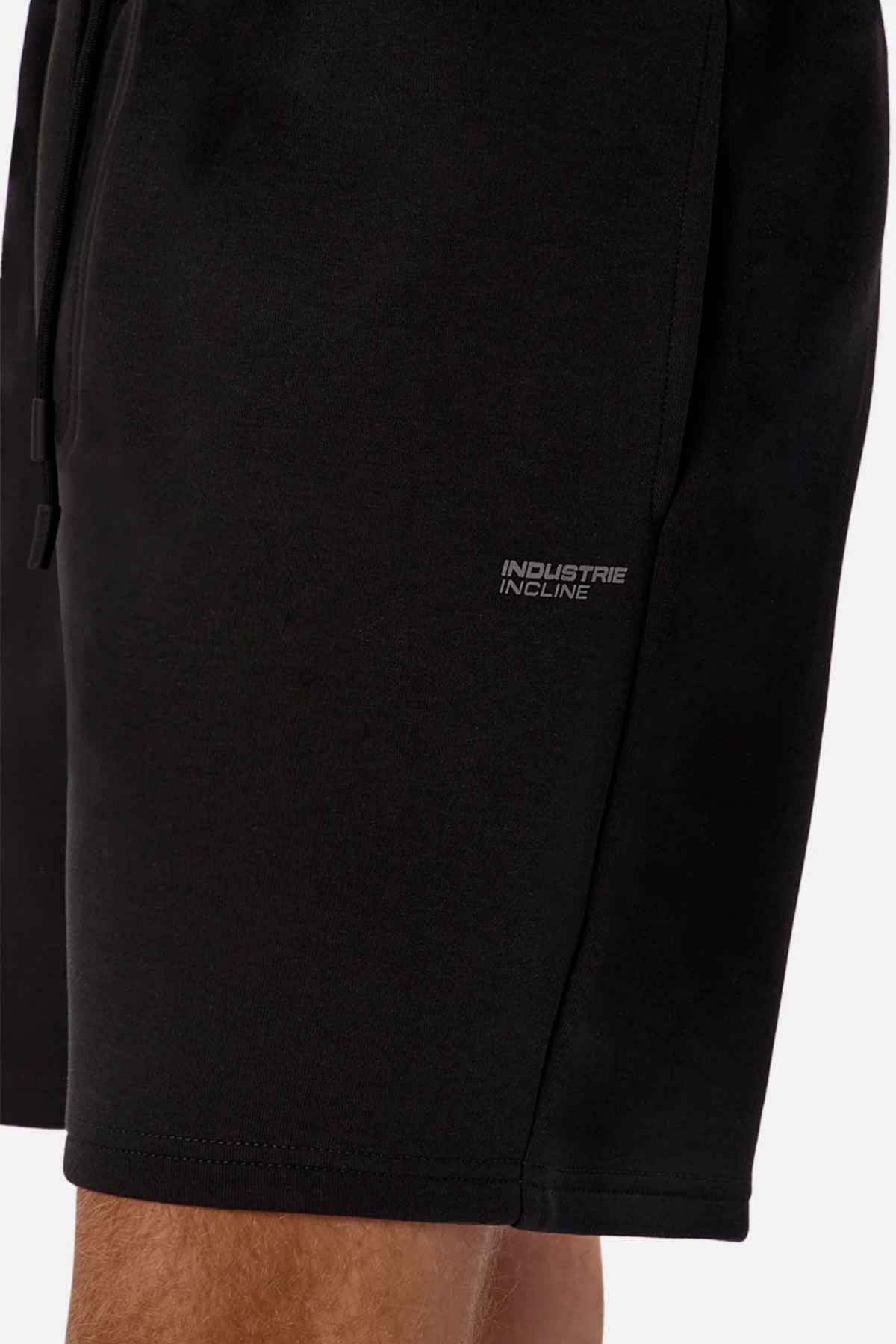 Close-up of a person wearing Industrie The Tech Track Short in black, showcasing "INDUSTRIE INCLINE" printed on the side. These shorts are crafted from soft, athletic material and include an elasticated drawstring waist. The background is plain white.