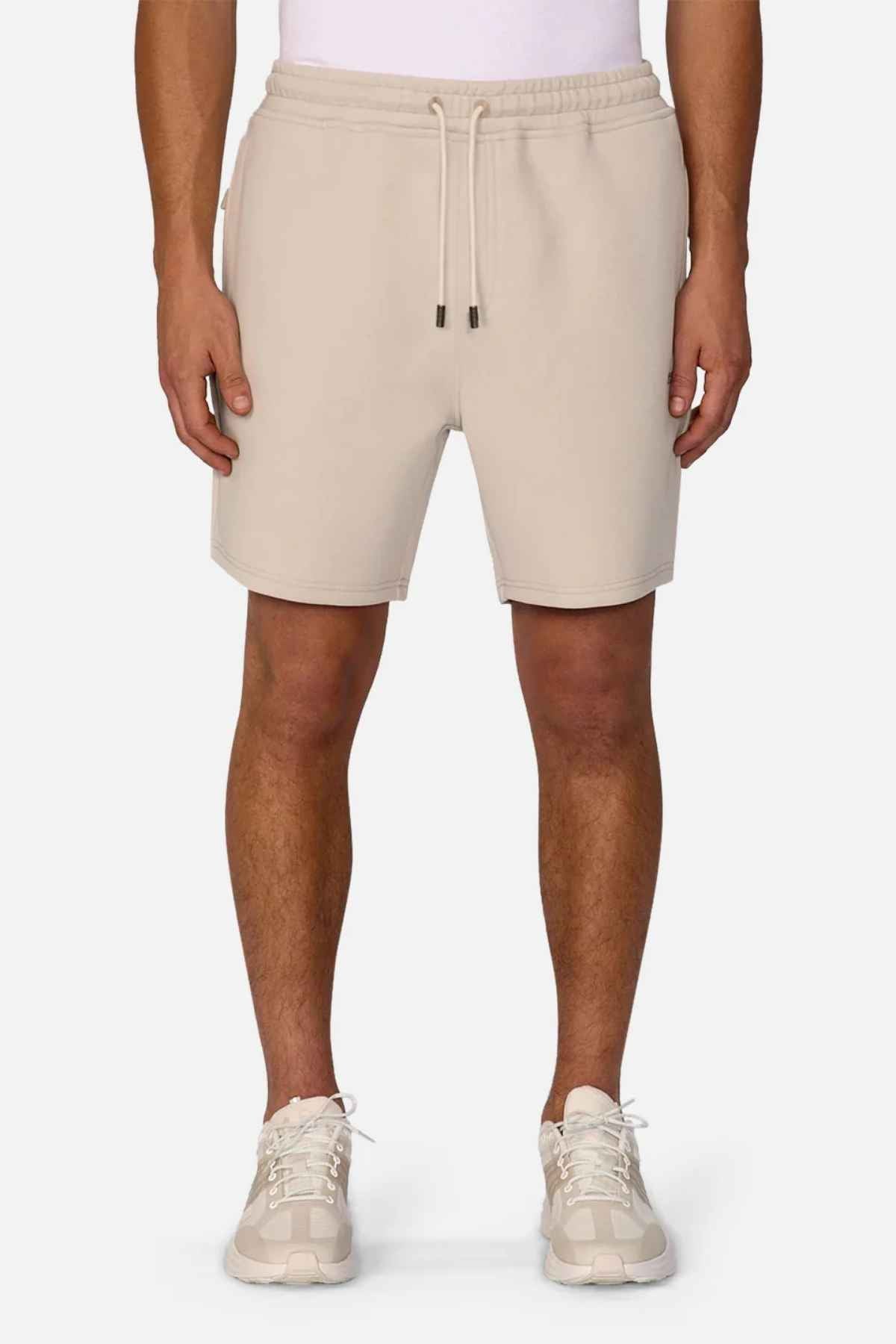 A person is wearing the Industrie The Tech Track Short in beige, featuring athletic material and a drawstring waist, paired with a white top and light-colored sneakers. This versatile design is perfect for any occasion, set against a plain white background.