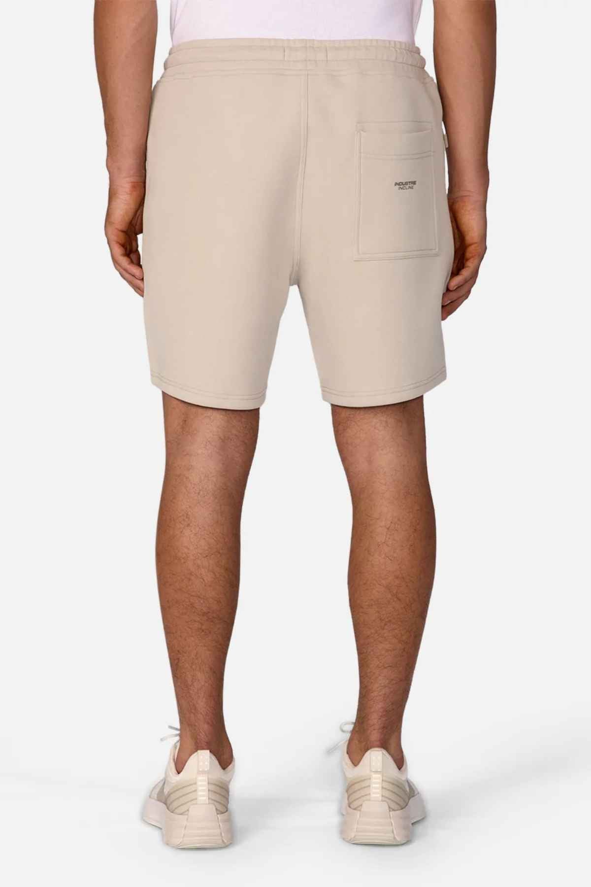 A person wearing The Tech Track Short by Industrie, featuring a versatile design in beige with the INCLINE logo on the back pocket, stands with white sneakers, facing away from the camera.