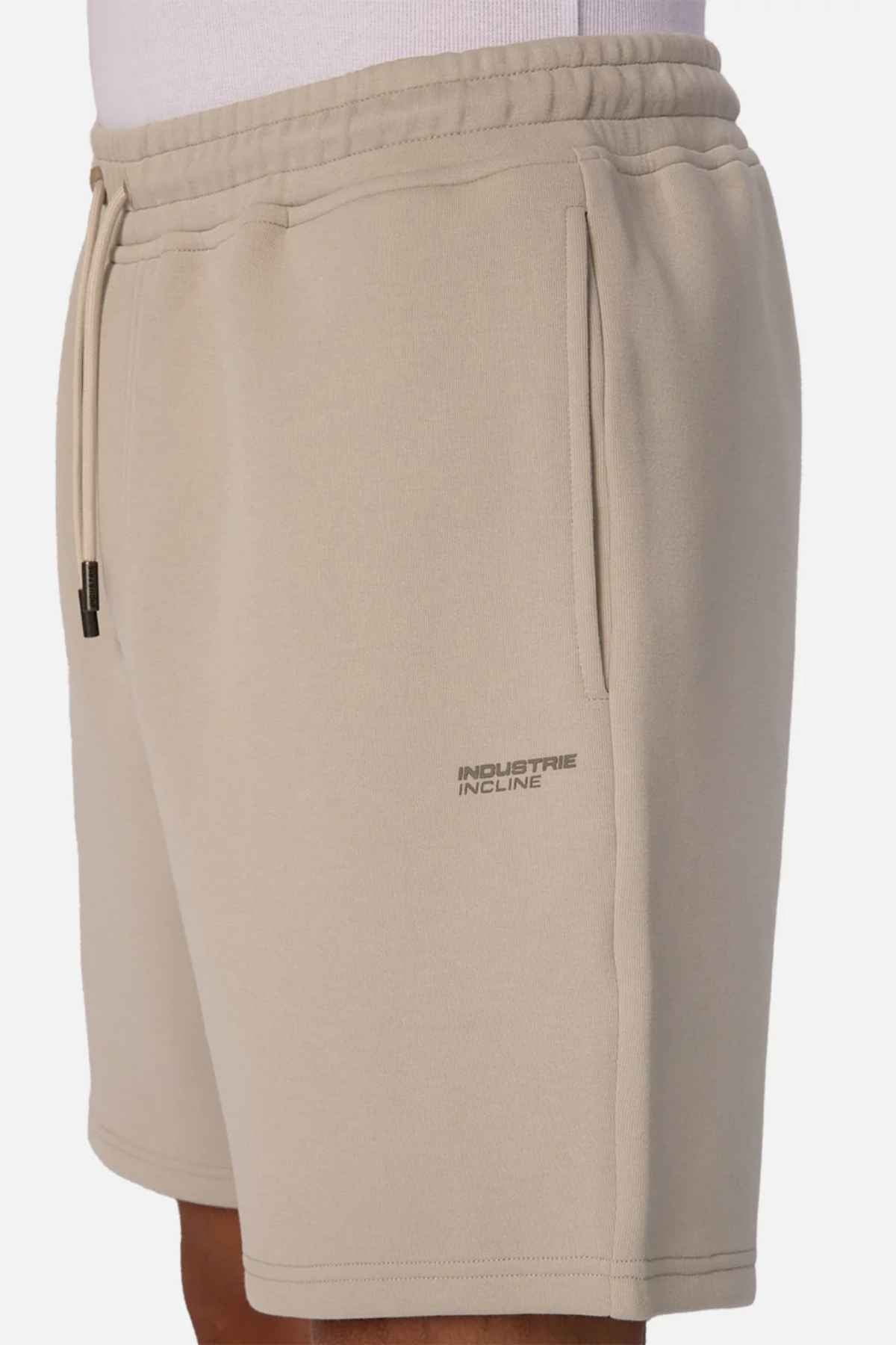 A person is wearing The Tech Track Short in beige, featuring a drawstring waist for adjustable comfort. These shorts, crafted from athletic material, display a compact "Industrie INCLINE" logo on the left side. The dynamic and versatile design is showcased with a white shirt, highlighting the side view of these sporty shorts.