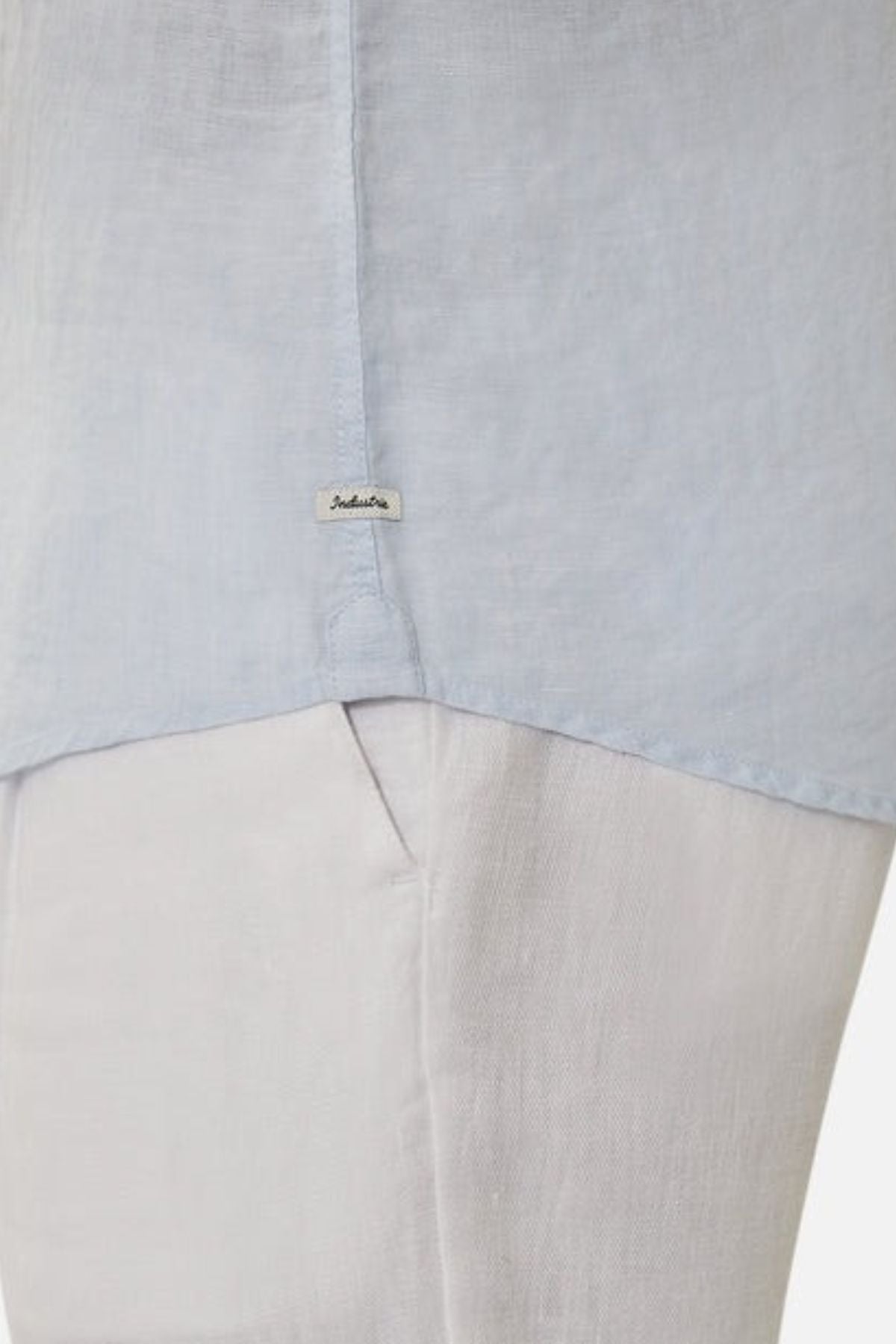 Close-up of a person wearing the Industrie The Tennyson Linen Blend Shirt in light blue, paired with white pants. The focus is on the shirt's lightweight fabric texture and a small label near the hem, showcasing fine details in the weave.