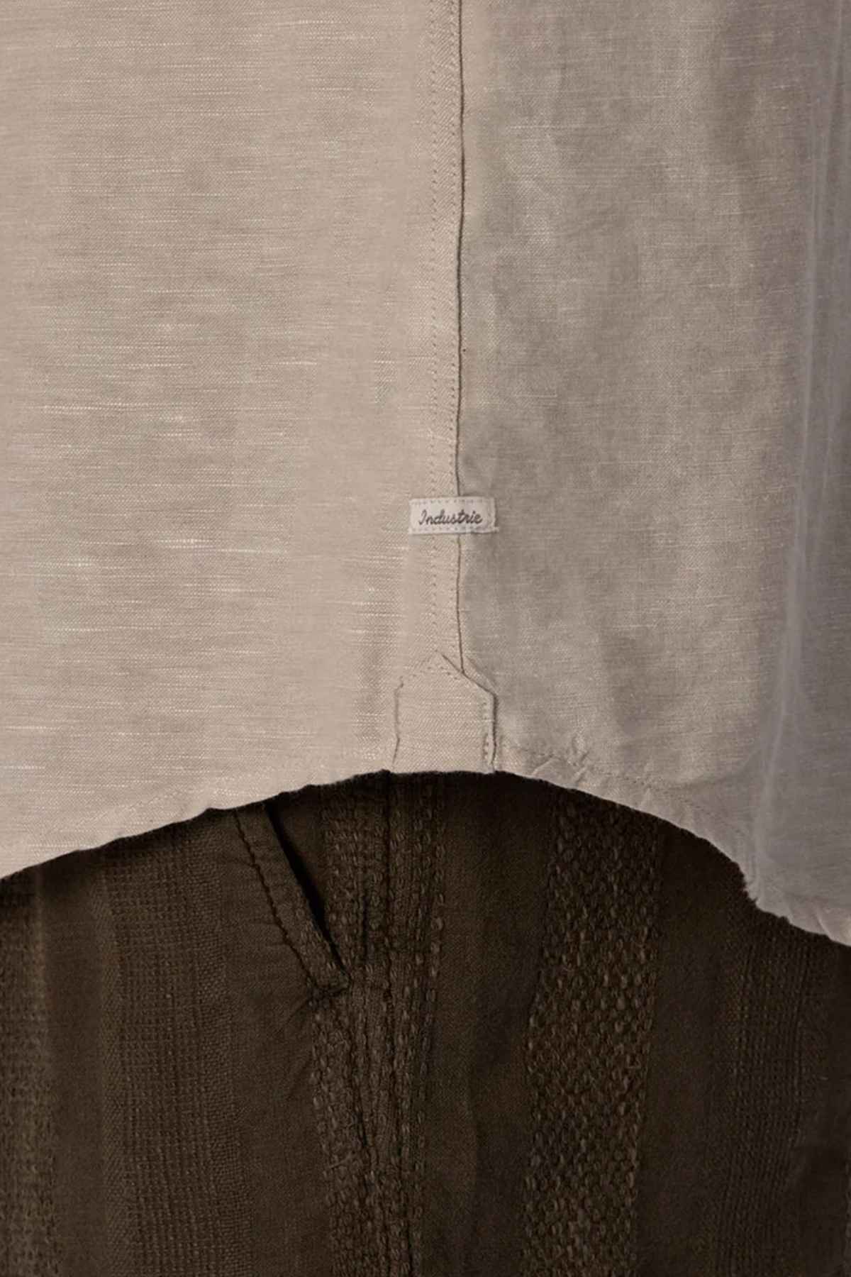 Close-up of a gray fabric with a small tag reading "Indelible," above dark textured pants. The image highlights the clothing materials and stitching details, reminiscent of a finely crafted Industrie The Tennyson Linen Blend Shirt, perfect for those breezy days.