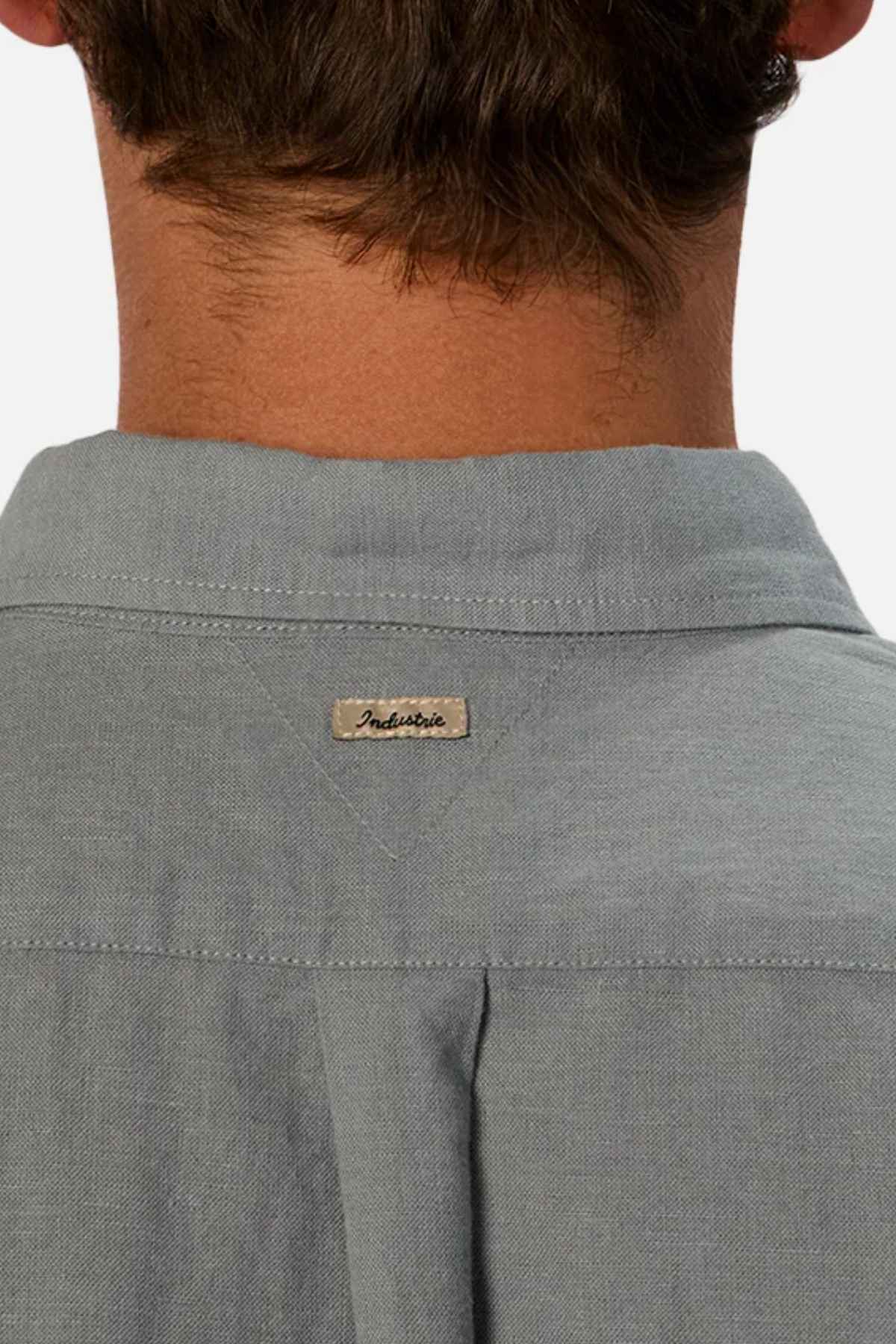 A close-up of the back of a person's neck wearing the Industrie The Tennyson Linen Blend Short Sleeve Shirt highlights its summertime comfort and versatility, thanks to its light gray color. The brand name "Industrie" is sewn on a small label below the collar, complementing the person's dark, short hair.