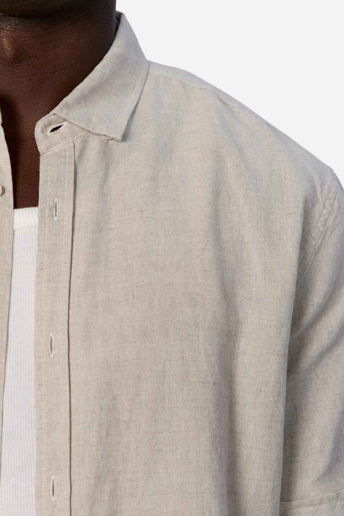 Close-up of a person's upper body, emphasizing summertime comfort in the Industrie The Tennyson Linen Blend Short Sleeve Shirt in light beige, unbuttoned and layered over a white ribbed knit shirt. This versatile shirt accentuates the person’s neck and shoulder against a plain background.