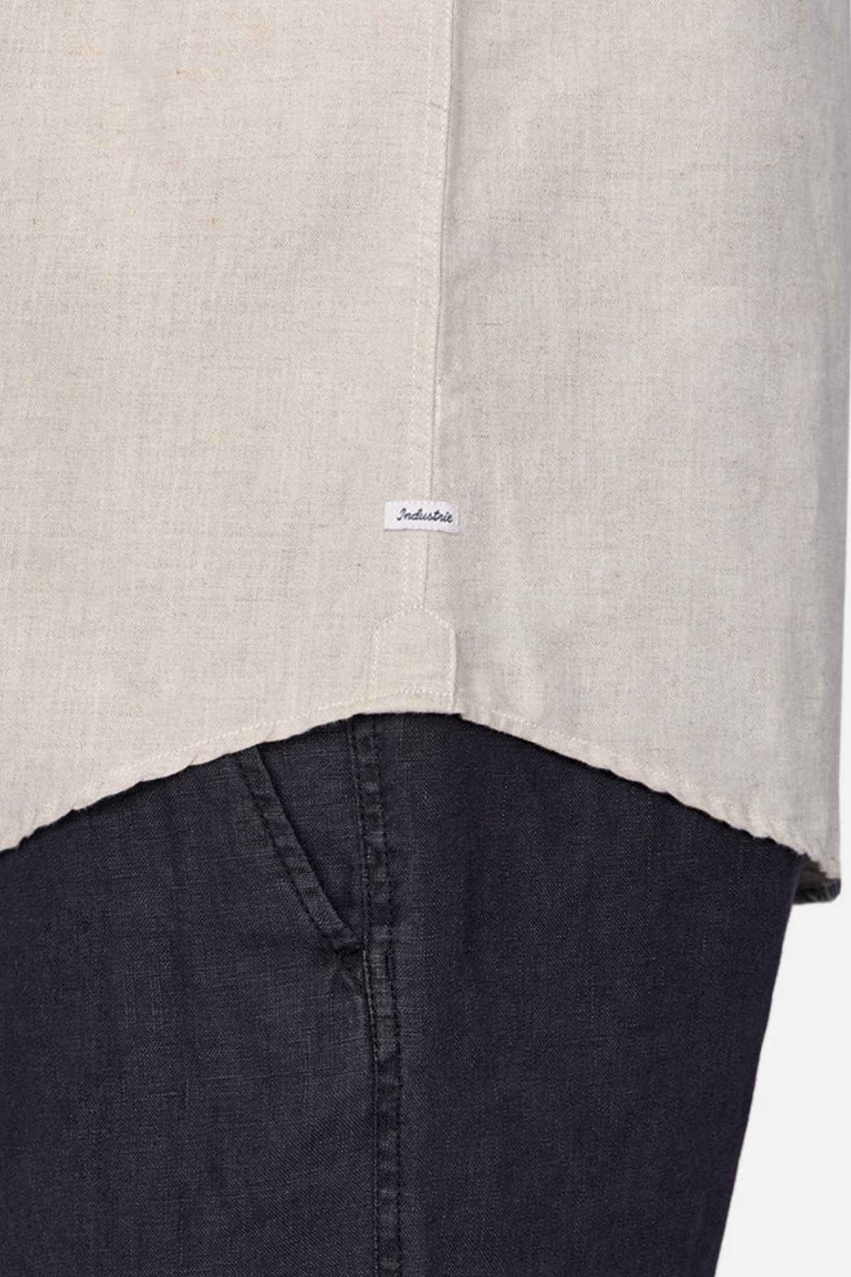 Close-up of a person wearing the Industrie The Tennyson Linen Blend Short Sleeve Shirt in versatile light beige, paired with dark blue pants. The shirt, ideal for summertime comfort, is adorned with a small label on its lower edge. Both garments appear to feature textured fabric.
