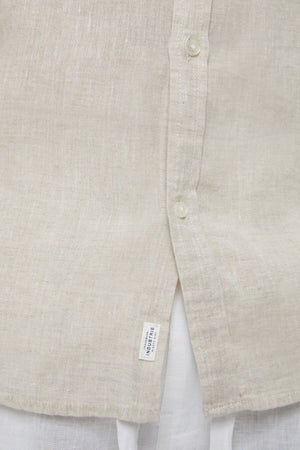 Close-up of the lower front of the Industrie The Tennyson Linen Short Sleeve Shirt in beige, showing two buttons, a blue-and-white tag near the hem, and white fabric underneath—emphasizing this classic shirt’s lightweight breathability.