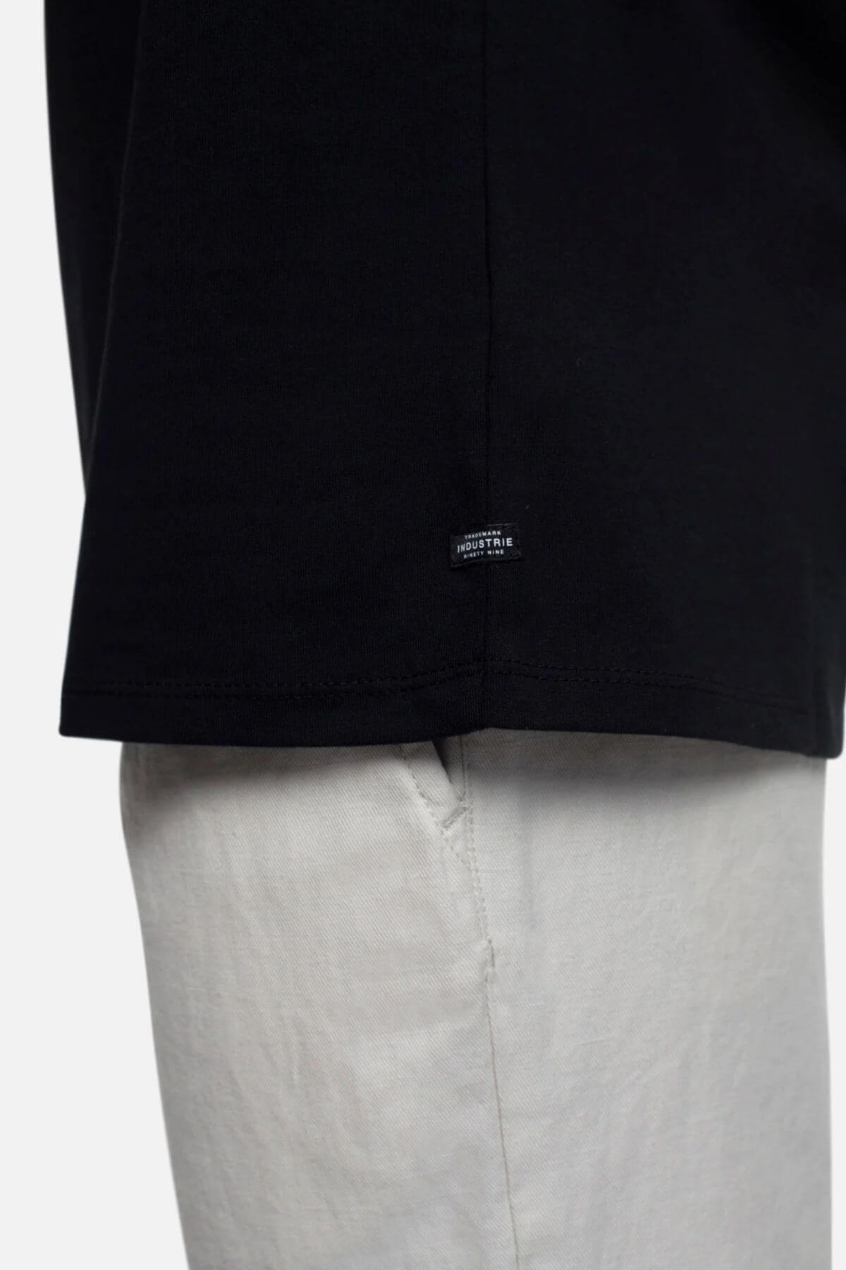 Close-up of a person wearing the Industrie The Triple G Sleeveless Tee in black, showcasing a relaxed fit with a small hem label, paired with light-colored pants. Focus is on the lower half of the shirt and the top section of the pants.