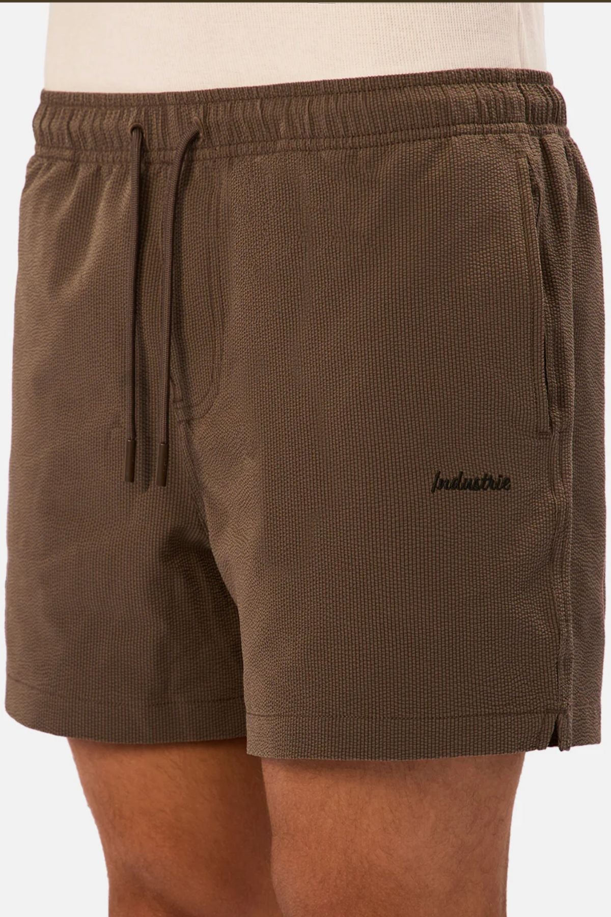 A person wearing Industrie The Tropez Swim Short in brown with textured fabric, elastic waistband, drawstring, side pockets, and "balusine" embroidered in black on the left thigh.