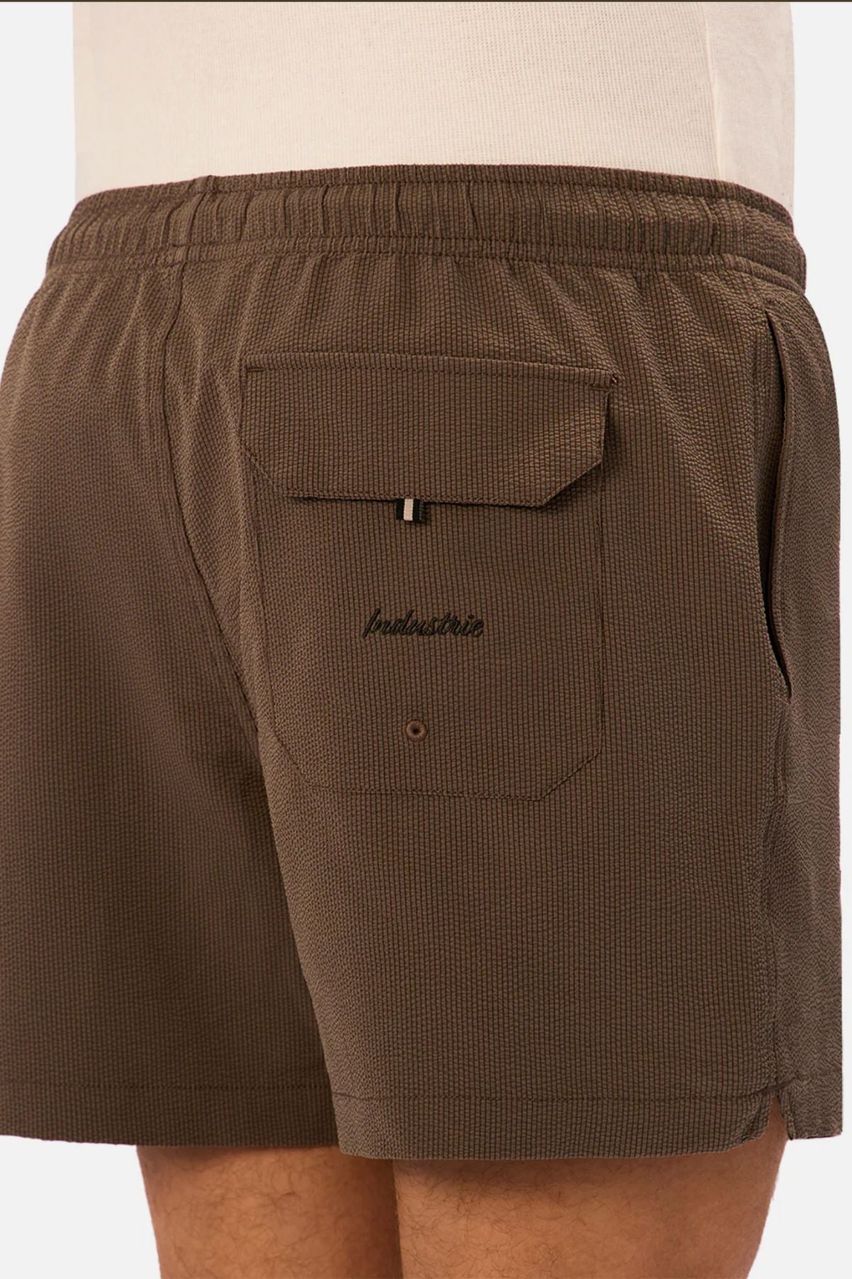 Close-up of a person wearing Industrie The Tropez Swim Short in brown, featuring textured fabric, elastic waistband, side pocket, back flap pocket with Industrie branding and "bankshot" embroidered above. Styled with a white top.