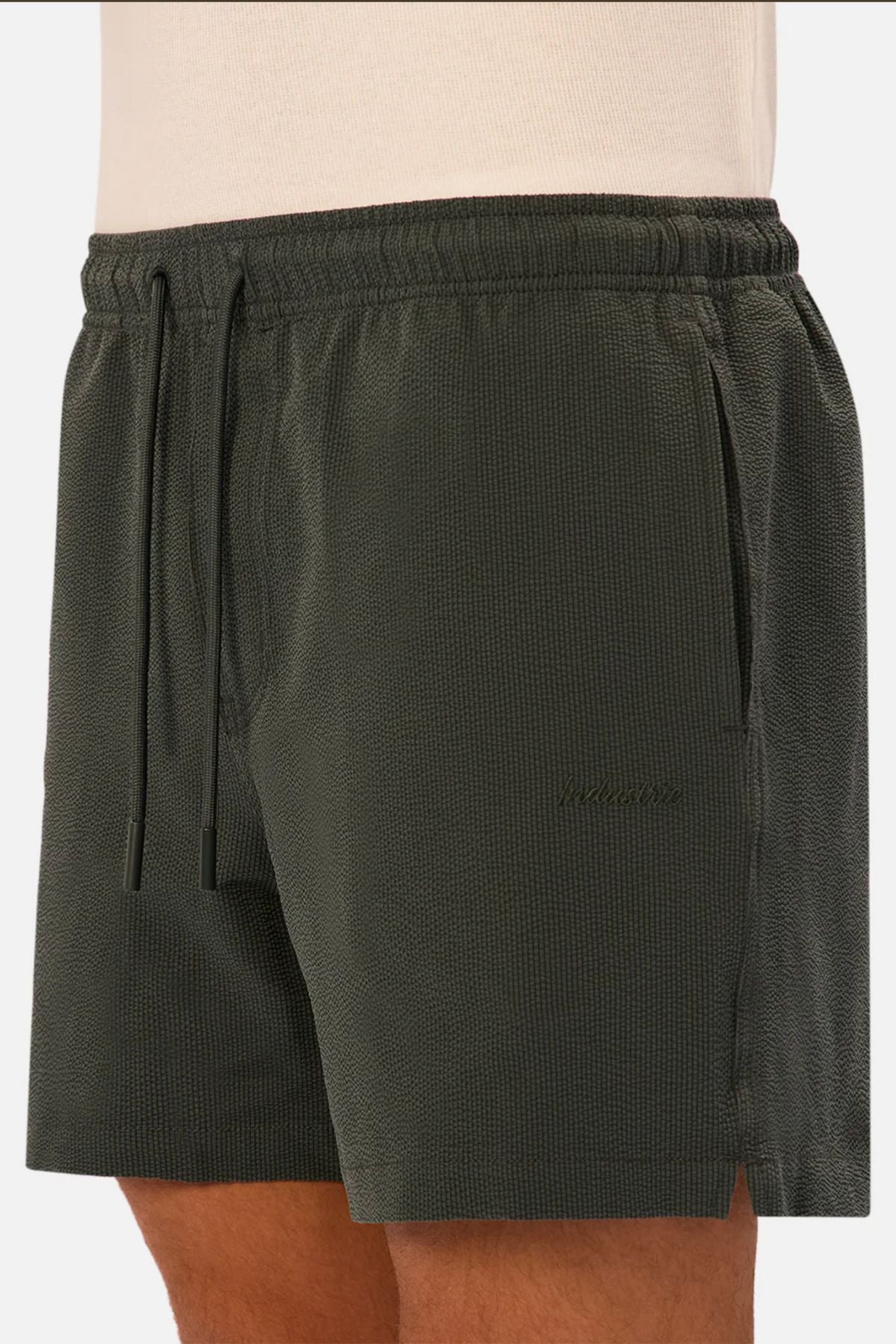 A person wears Industrie The Tropez Swim Short in dark green—elastic waist, side pockets, subtle left-leg embroidery, and drawstring—paired with a light beige top.