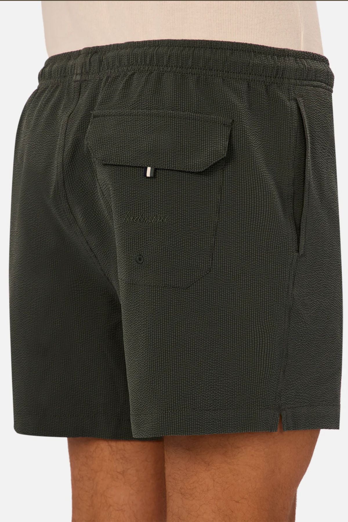 A close-up of a person wearing Industrie The Tropez Swim Short in dark green, featuring an elastic waistband, side pocket, and flap back pocket with loop detail. The model stands sideways to highlight these men's swimwear essentials.