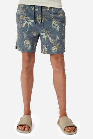 A person wears Industrie The Viloria Bahama Short—blue floral-patterned swim shorts with an elastic waistband—paired with a light shirt and beige slippers; only the lower torso and legs are shown against a plain background.