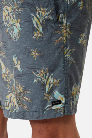 A close-up of a person's leg wearing Industrie The Viloria Bahama Short, featuring a dark gray tropical floral print in blue, yellow, and white. An elasticised waistband offers comfort, with a black "Industrie" label by the pocket seam.