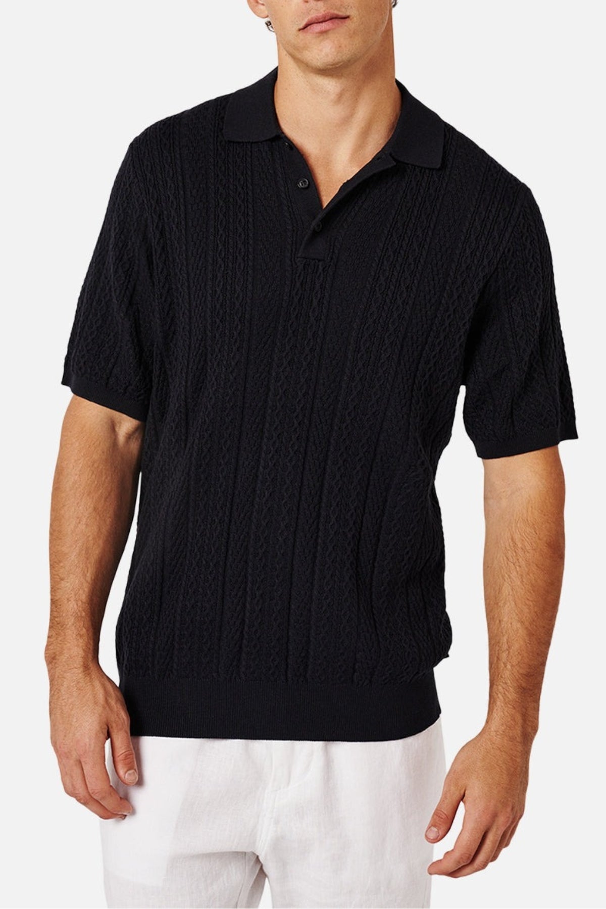 A person, face partially visible, wears the black Industrie The Vito Polo with a subtle cable knit finish and button collar. Paired with white pants, they stand against a plain white background.