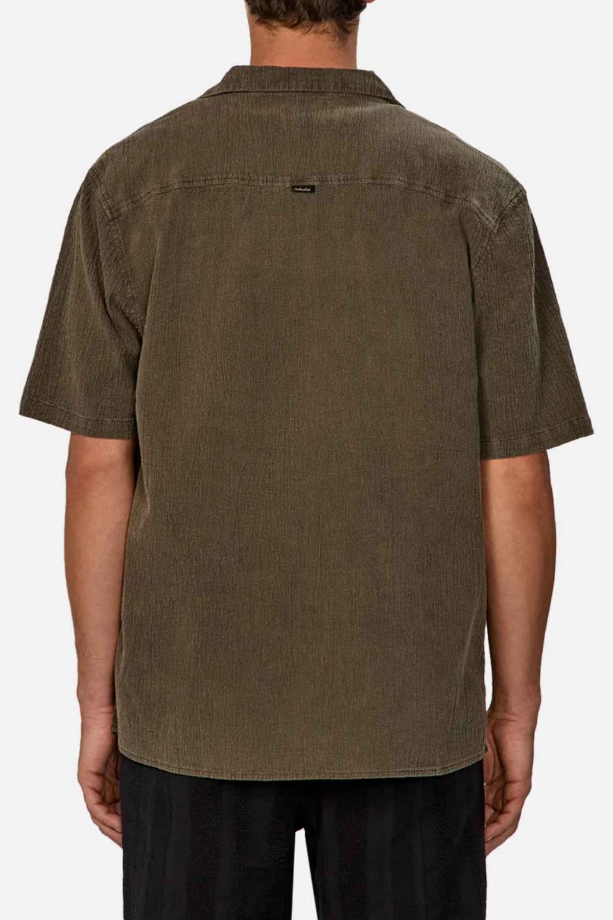 A person in the Industrie The Washed Magna Short Sleeve Shirt, with its olive green color and oversized fit, stands with their back to the camera. Made from breathable fabric and featuring a small label near the collar, it pairs well with dark pants against a plain white background.