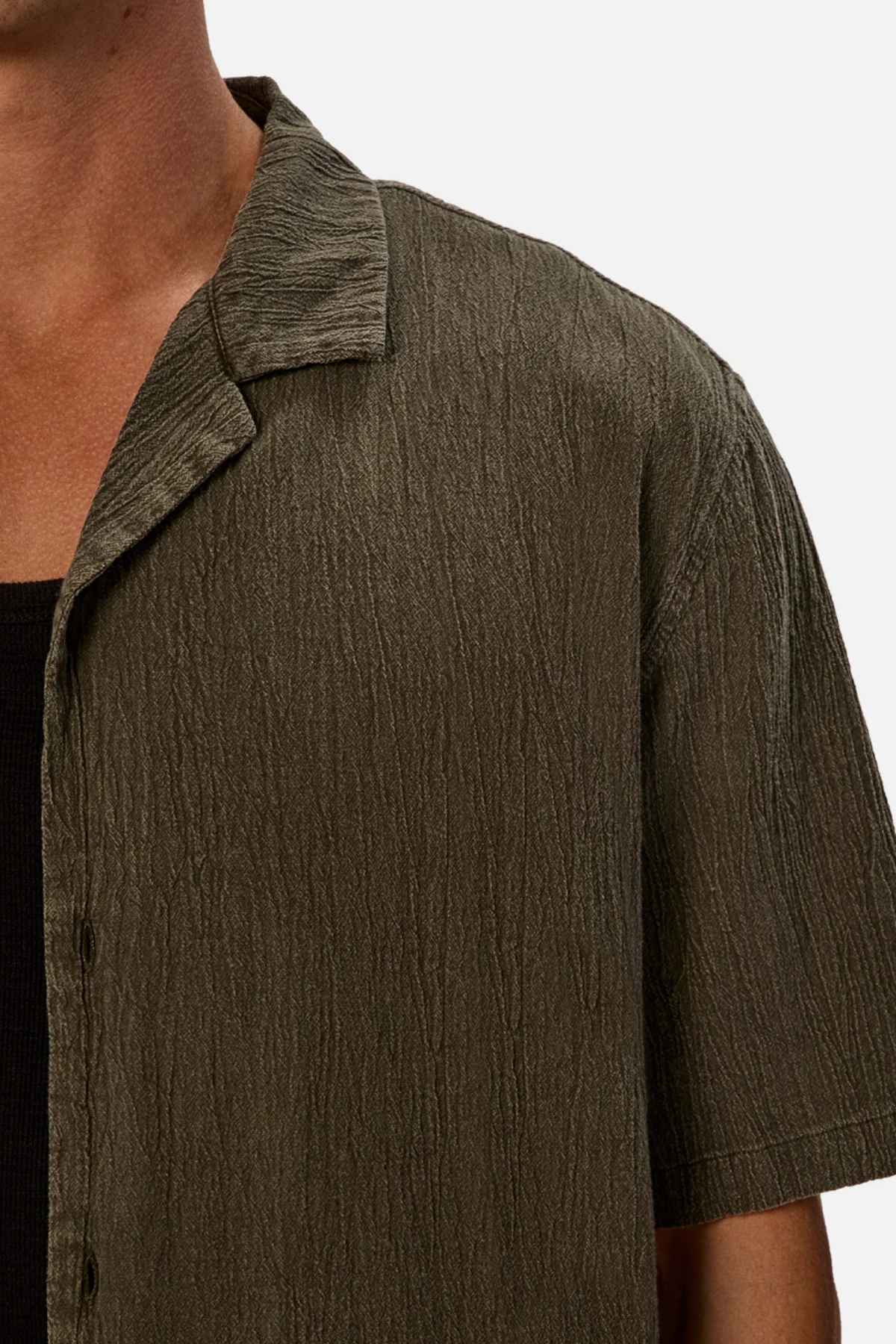A close-up captures a person in an oversized fit crinkled short-sleeve shirt in olive green, layered over a black top. This breathable fabric shirt, identified as the Industrie The Washed Magna Short Sleeve Shirt, is designed with a collar and buttons. The background is plain white.