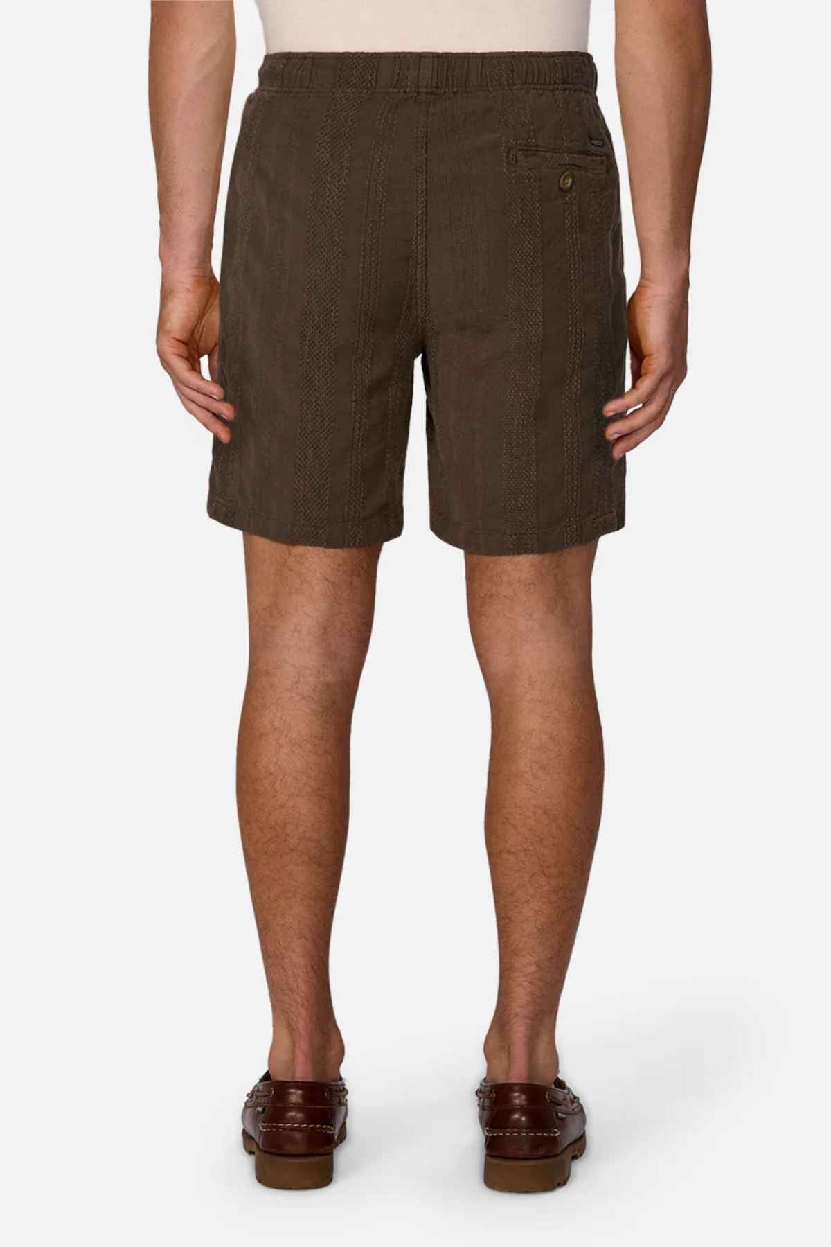 A person dressed in Industrie The Washed Salamanca Short and brown loafers is seen from behind. The textured cotton blend shorts showcase a tailored fit with a side pocket button, contrasting against a plain white background.