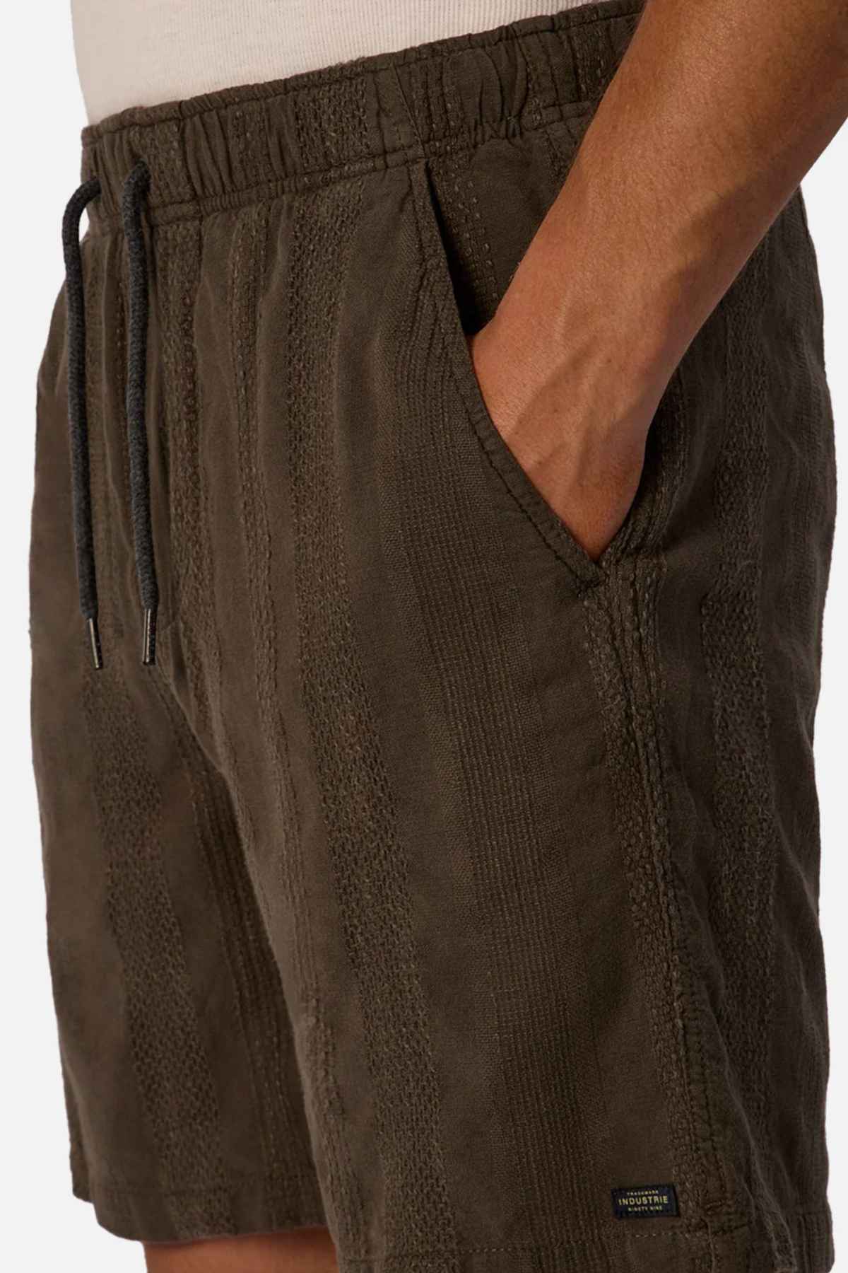A person wearing the dark green Industrie The Washed Salamanca Short made from a textured cotton blend stands with one hand casually resting in the pocket. These shorts boast a drawstring waistband, tailored fit, and a small black label near the hem, all set against a plain, light-colored background.