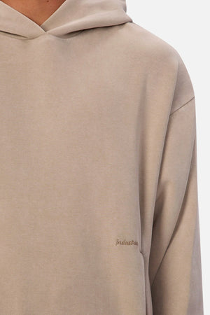 A close-up shows a person wearing the Industrie The Washed Tech Hood in light beige with a small embroidered "Industrie" logo on the chest. Only the upper torso and part of the hood are visible, highlighting its modern, casual style.