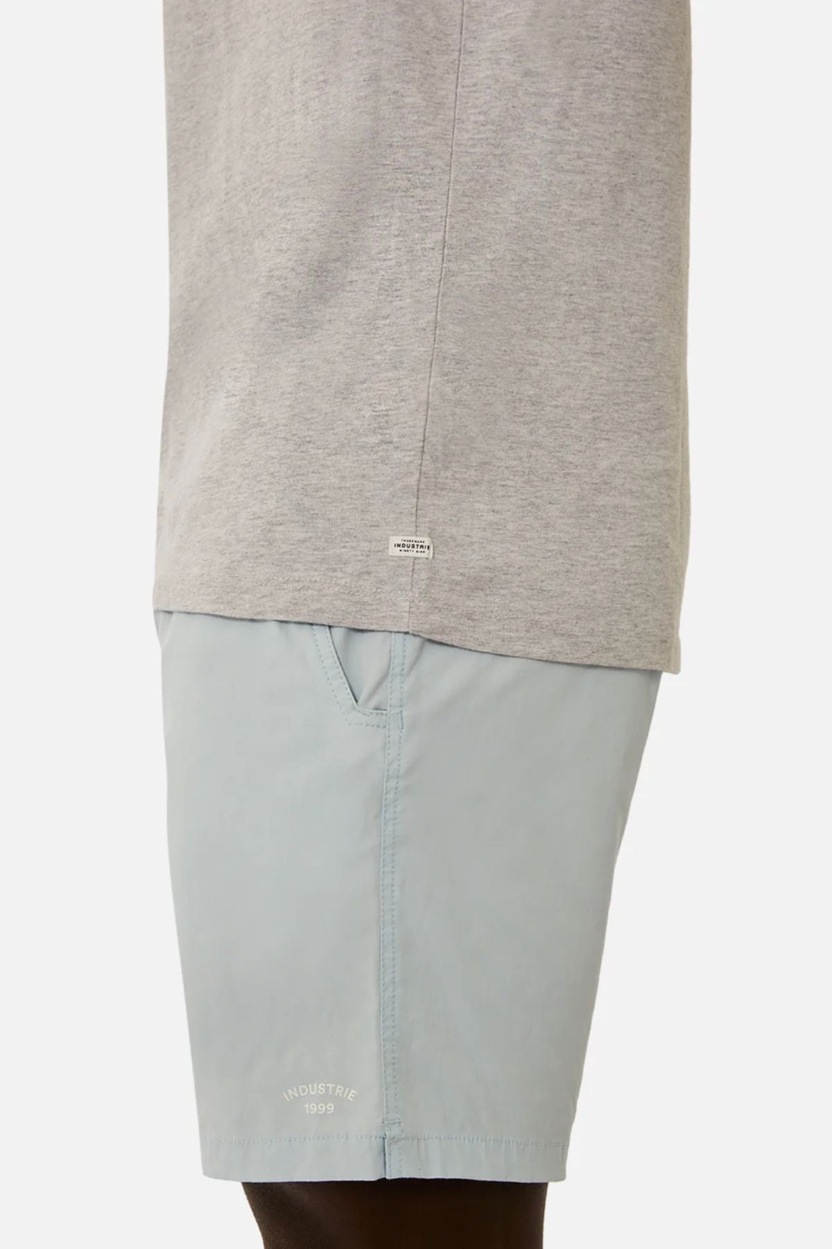 A person wearing a light gray Industrie The Triple G Sleeveless Tee, showcasing a relaxed fit and a small label near the hem, paired with light blue shorts with side pockets, stands against a plain background.