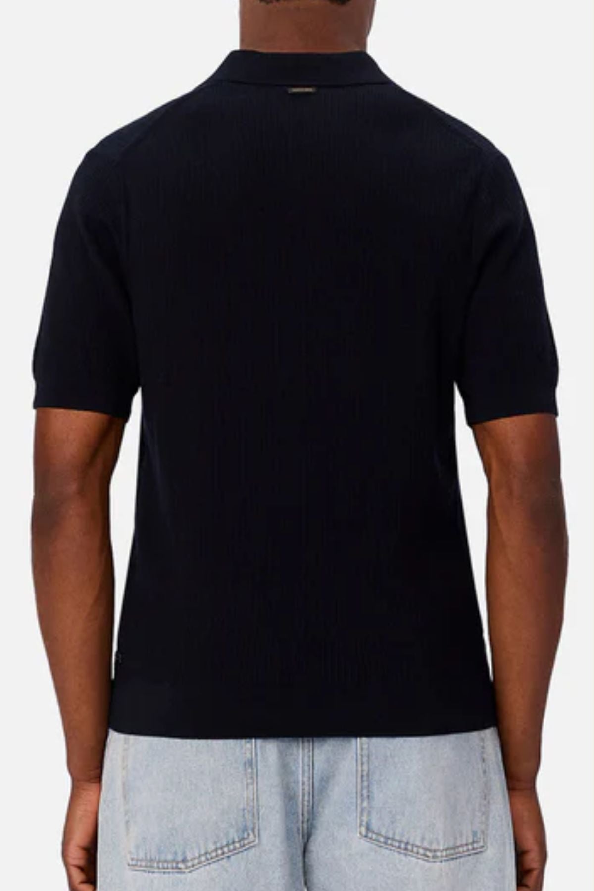 A person wearing the Industrie The Valencia Polo, a short-sleeve black 100% cotton shirt, with light blue jeans, shown from the back against a plain light background.