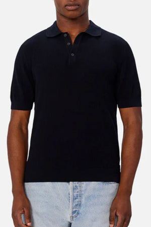 Someone wears the Industrie The Valencia Polo, a short-sleeved black lightweight 100% cotton knit with collar buttons, paired with light blue jeans and stands against a plain light background.