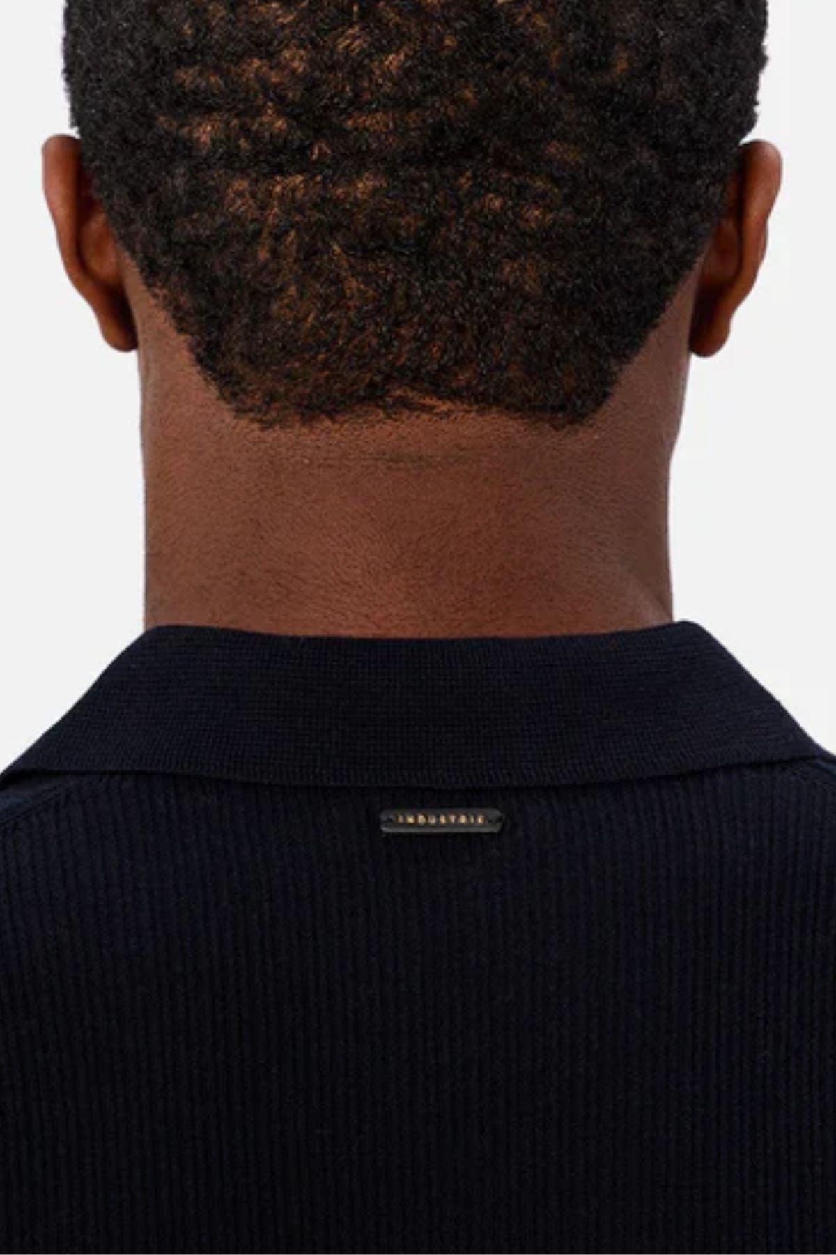 A close-up of the back of a person’s head with short curly hair, wearing the Industrie The Valencia Polo—a dark, lightweight 100% cotton knit featuring a small rectangular collar label.
